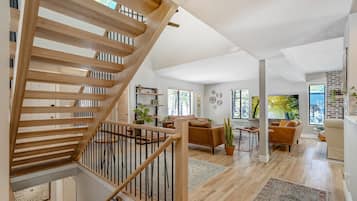Uma sala de estar ampla com escada de madeira, conjunto de sofás e uma grande janela com vista para o exterior.