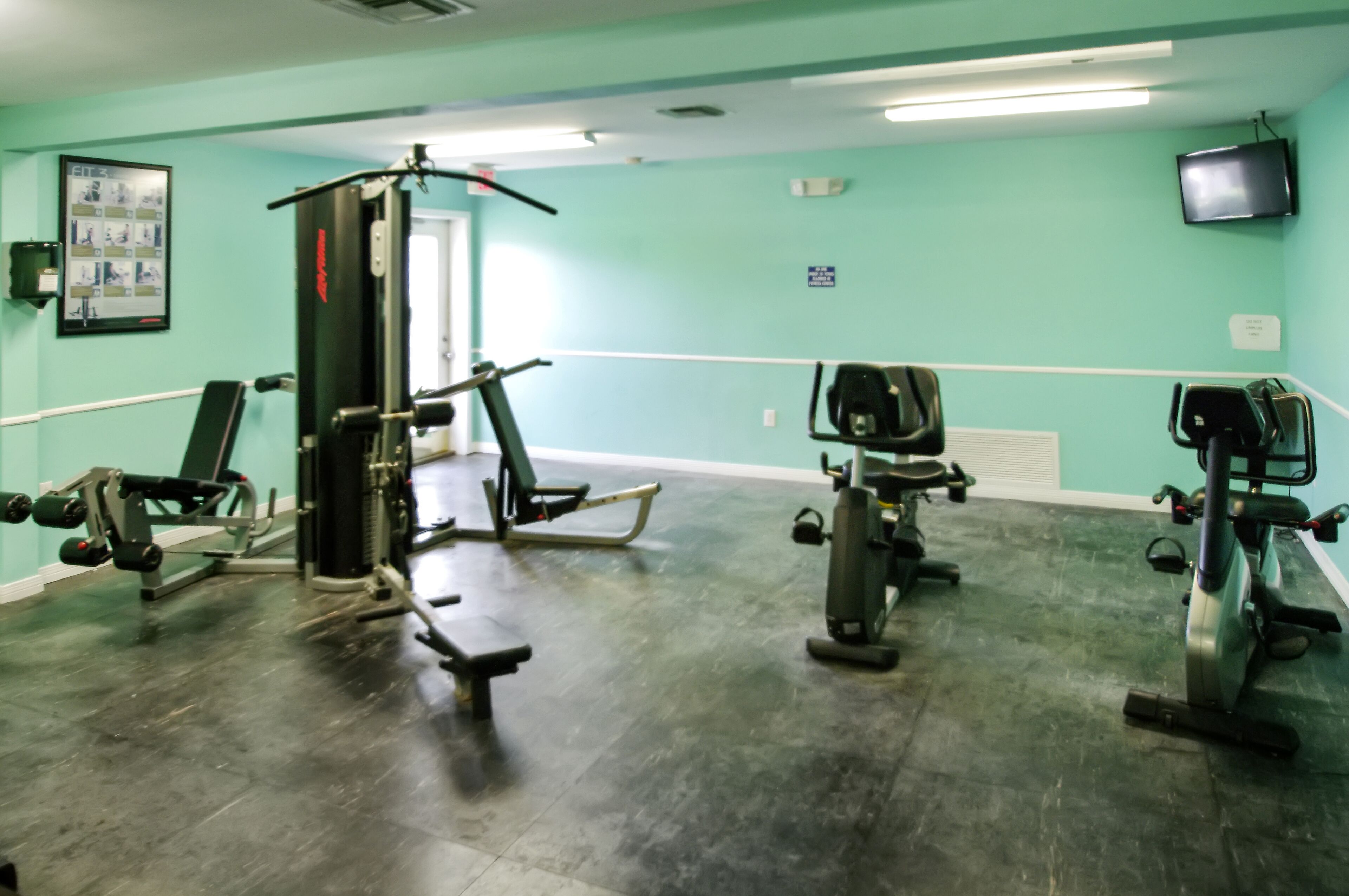 Een sportschool met diverse fitnessapparaten, teal muren en een televisie aan de muur.