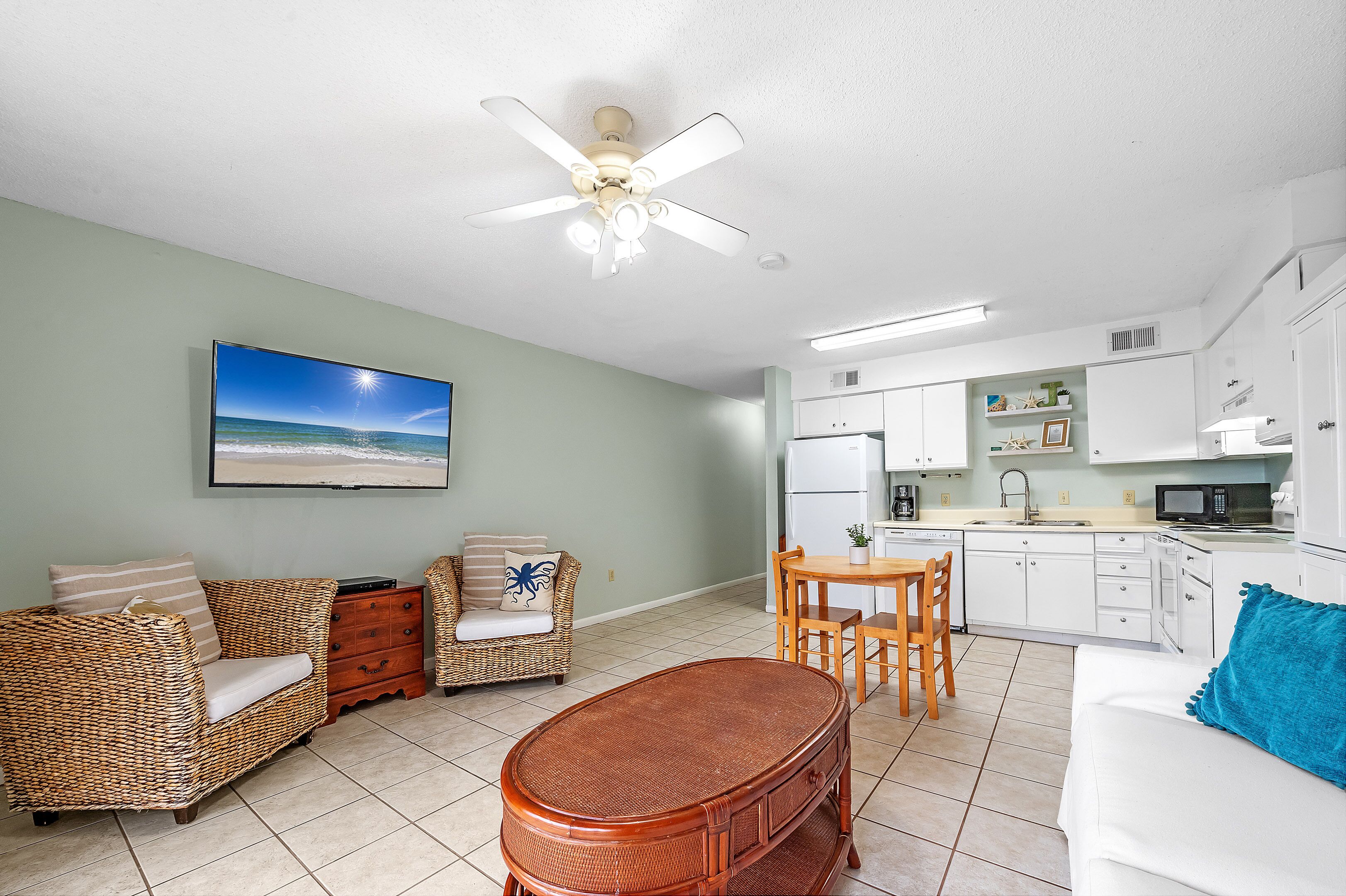 A living room with a television, wicker furniture, a ceiling fan, and a kitchen area with white cabinets and appliances.