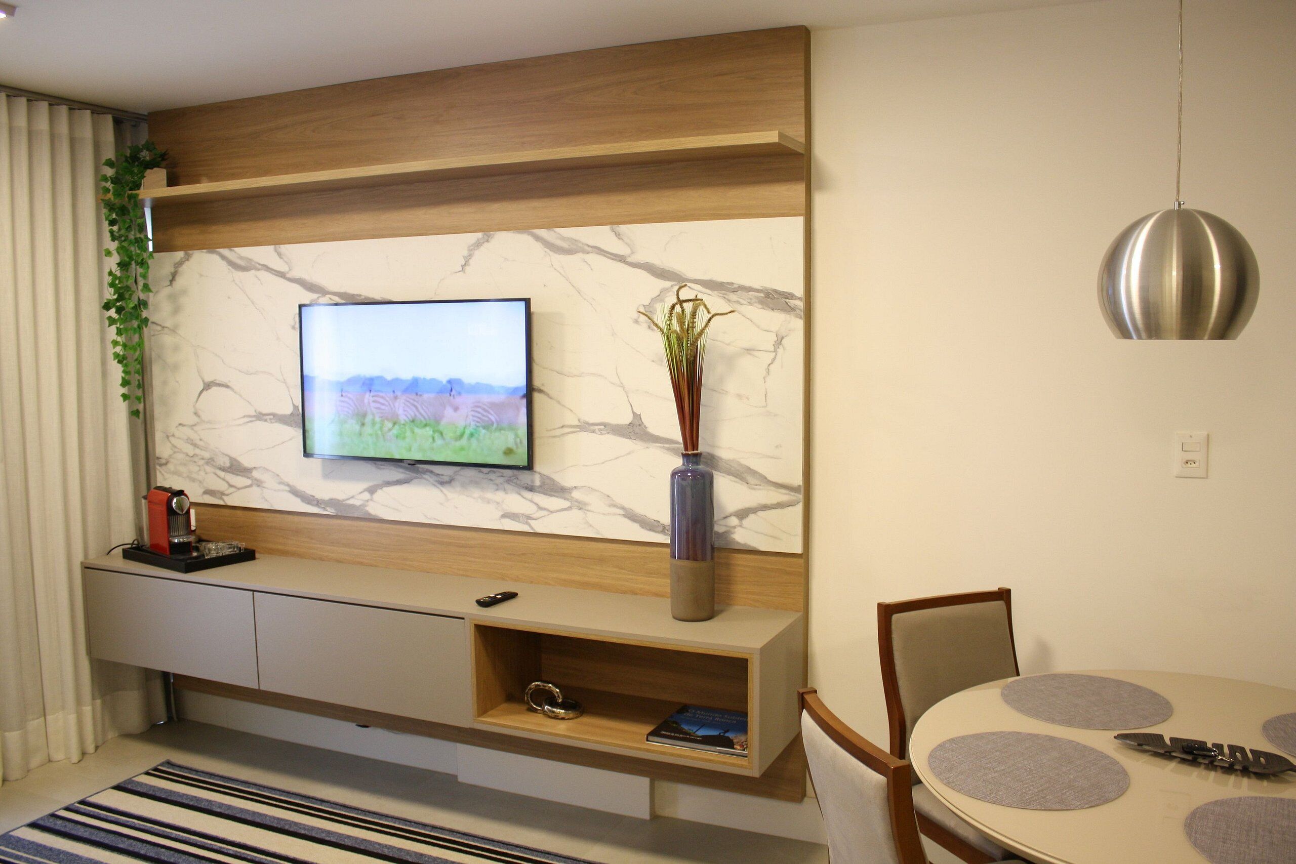 Een moderne woonkamer met een flatscreen televisie aan een marmeren muur, een houten console met een vaas, en een eethoek met een ronde tafel en stoelen.