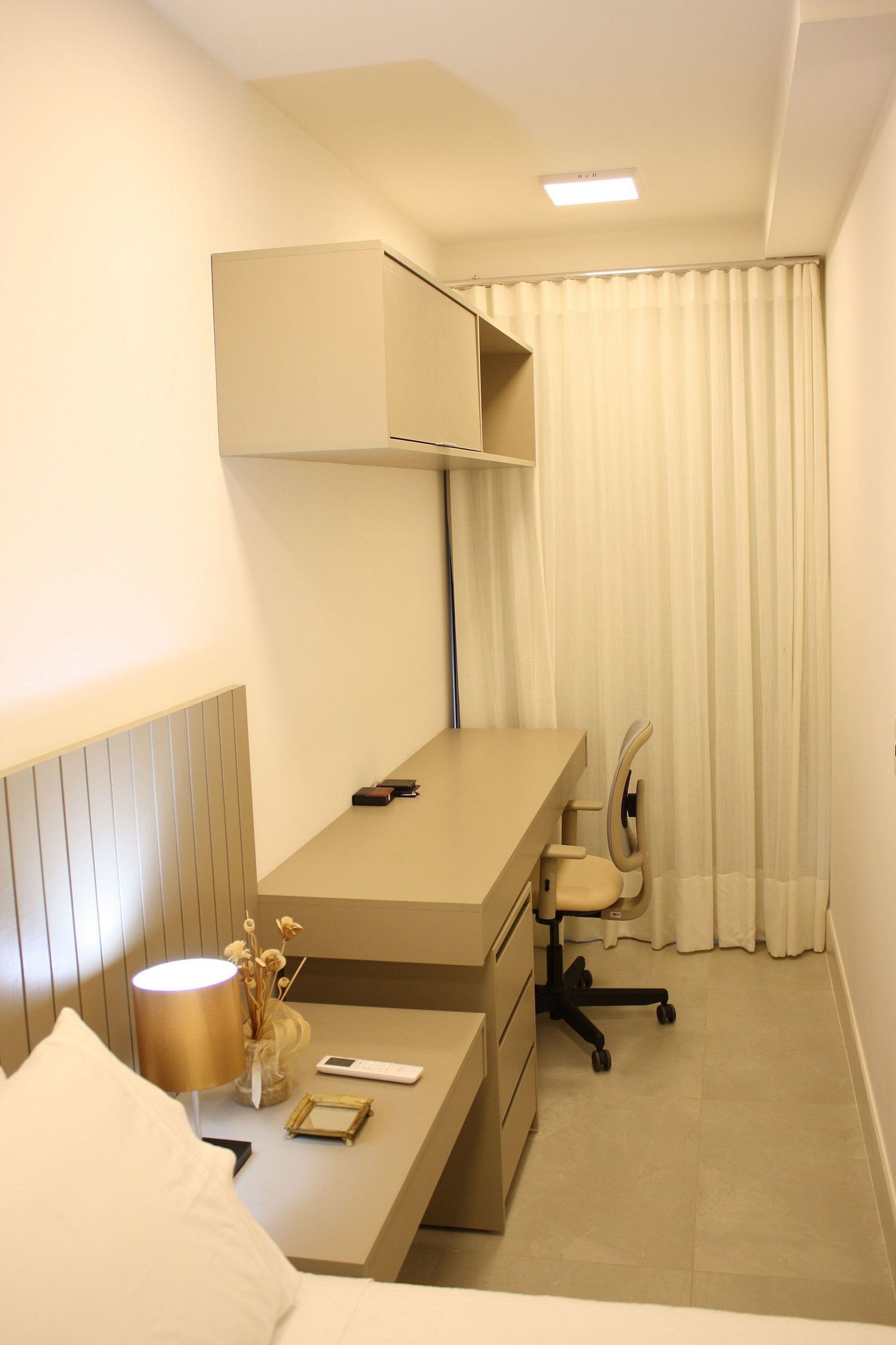 A hotel room with a bed, a desk with a chair, a lamp, a remote control, and a curtain.