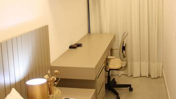 A hotel room with a bed, a desk with a chair, a lamp, a remote control, and a curtain.