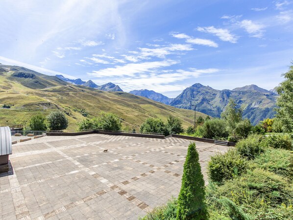 A courtyard with a tiled floor, surrounded by greenery and mountains in the background.