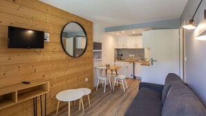 Ein modernes Wohnzimmer mit grauem Sofa, Holzboden, einem an einer Holzwand montierten Fernseher, einem kleinen Esstisch mit Stühlen und einer Küchenzeile mit weißen Schränken und einem Geschirrspüler.