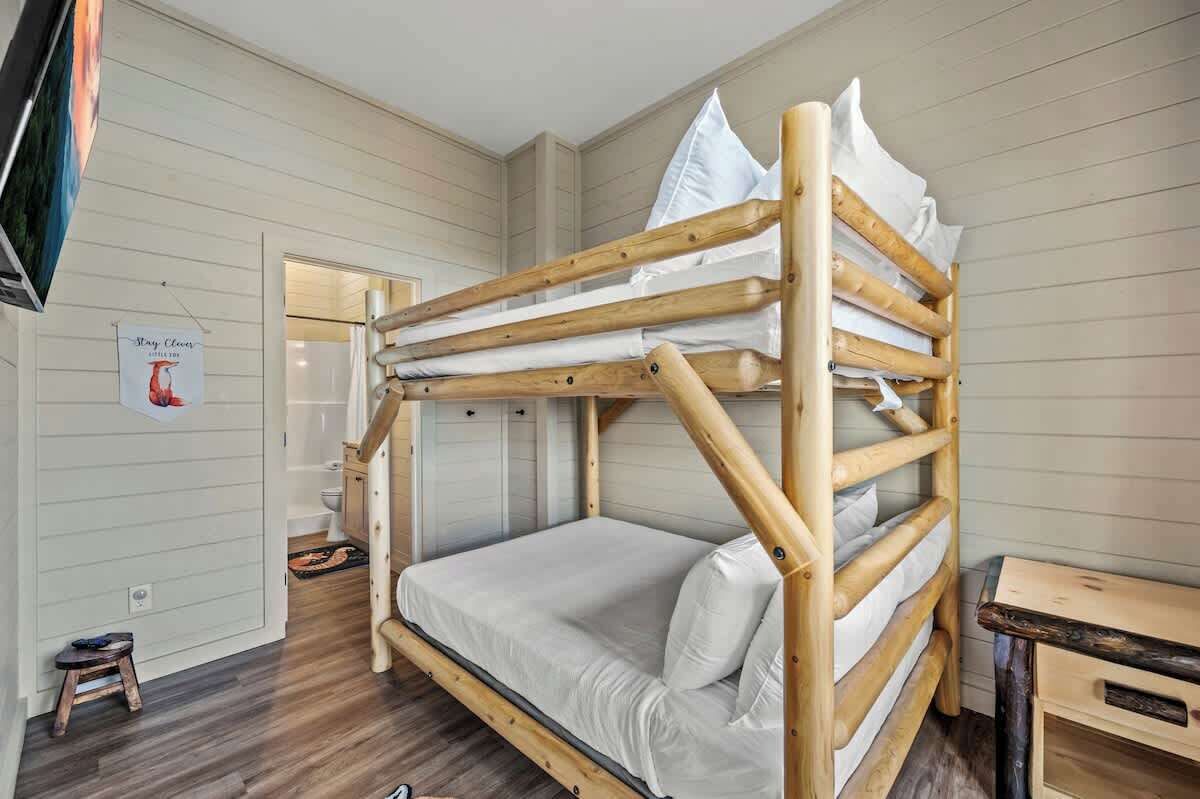 Un lit superposé avec une structure en bois et une literie blanche, un petit tabouret en bois et un téléviseur fixé au mur.