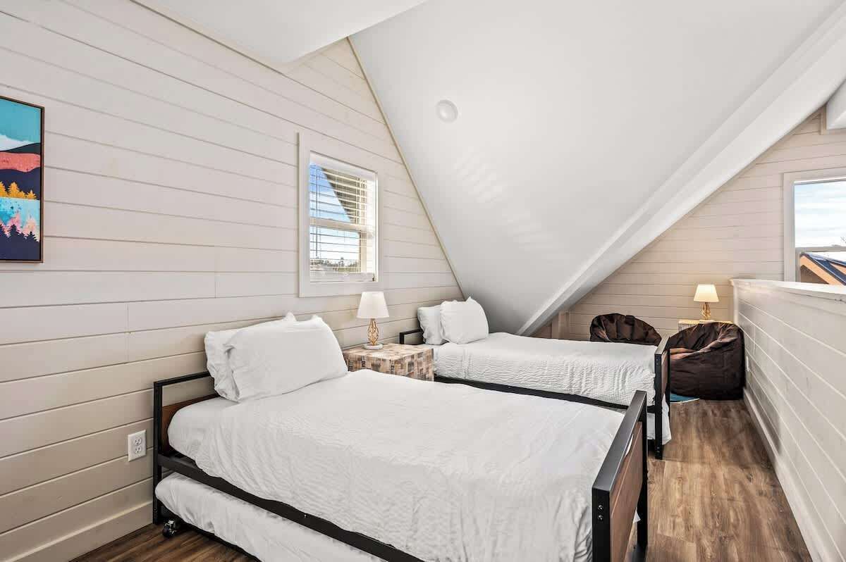 A bedroom with two beds, a window, a painting, and a small table with a lamp.