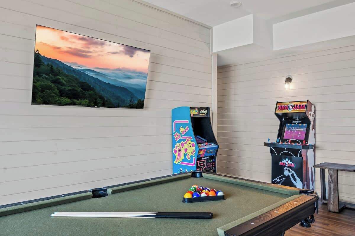 Una mesa de billar con bolas, una taco de billar, un televisor montado en la pared y máquinas de videojuegos.