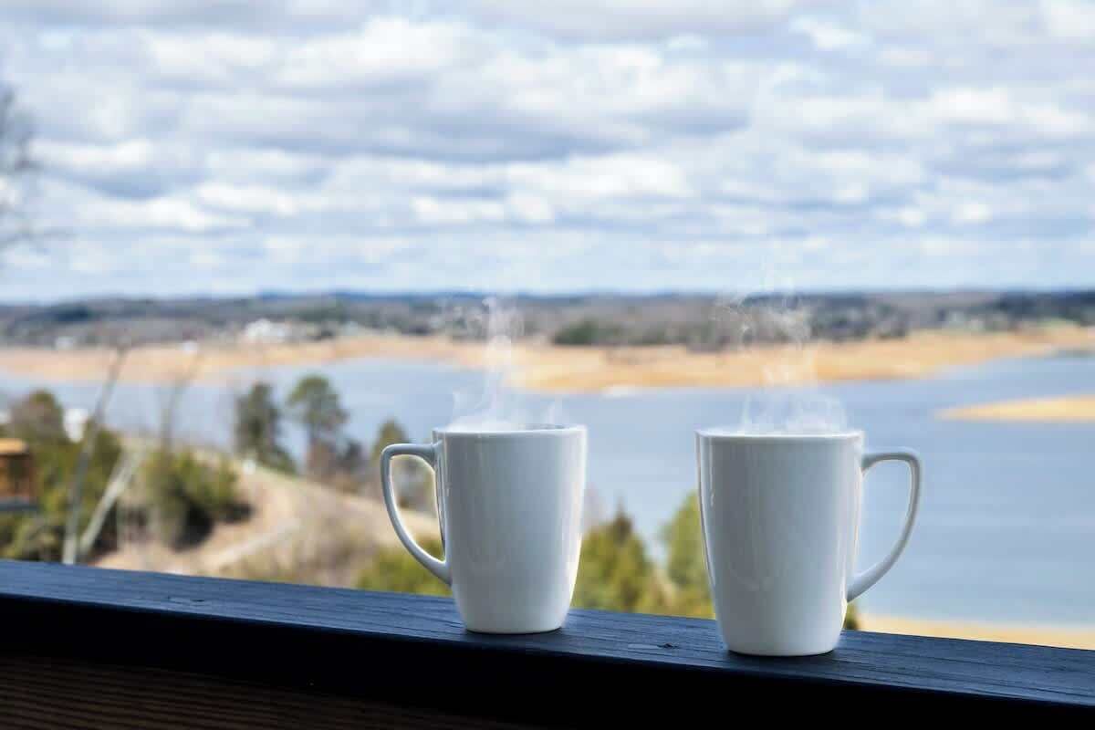 Two white mugs on a ledge with a view of a body of water and distant land.