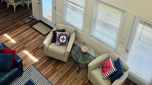 Amplia sala de estar con techo alto, ventilador de techo y vista al océano a través de grandes ventanas. Hay dos sofás con almohadones de temática náutica, una mesa de centro de vidrio y un comedor con mesa y sillas.