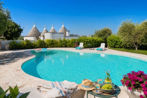 A swimming pool with lounge chairs, a table with drinks and snacks, and a building with conical roofs in the background.