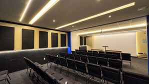Un salón de conferencias con filas de sillas, un atril, una pantalla de proyección y paneles acústicos en las paredes.
