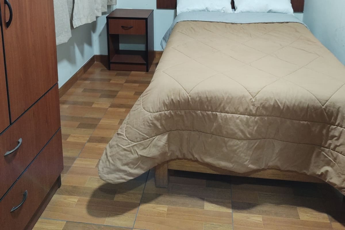 Une chambre à coucher avec une armoire en bois, une table de chevet, un lit recouvert d’une couette beige et une fenêtre avec des rideaux.