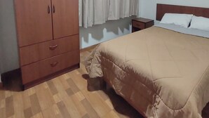 Une chambre à coucher avec une armoire en bois, un lit recouvert d’une couvre-lit beige, une table de chevet et une fenêtre avec des rideaux.