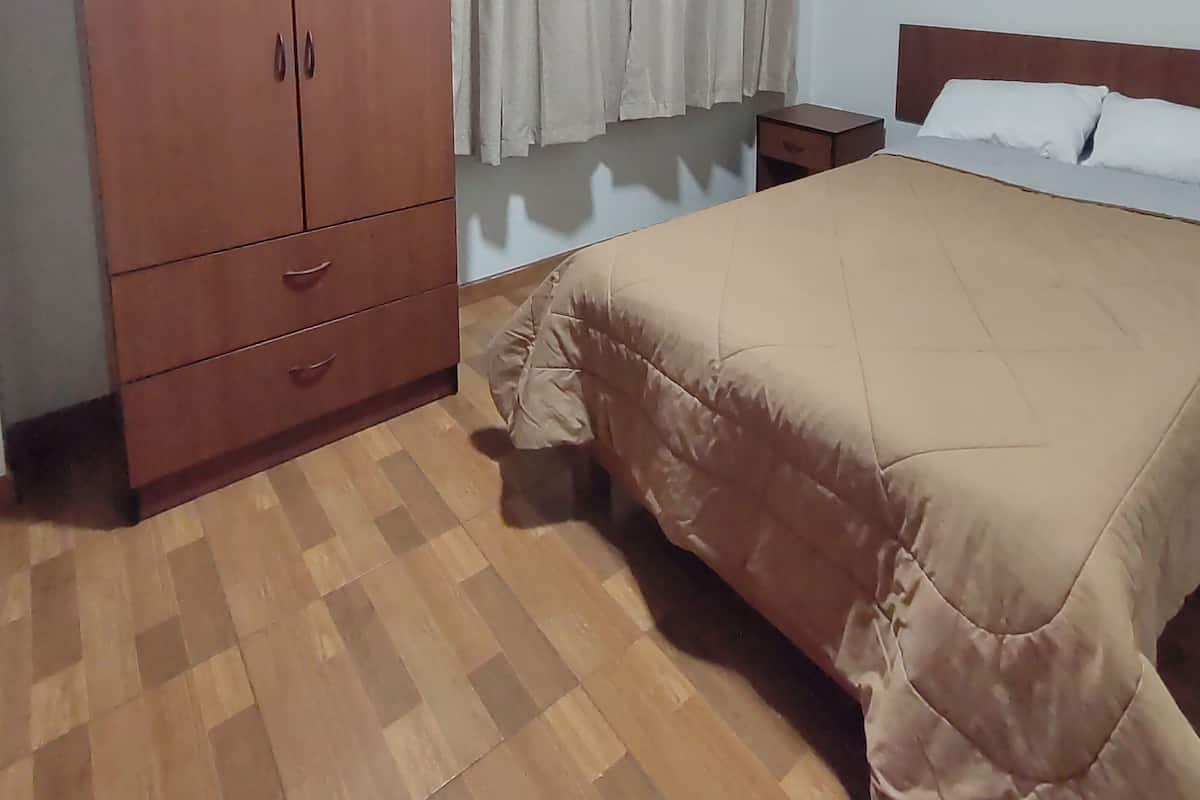 Une chambre à coucher avec une armoire en bois, un lit recouvert d’une couvre-lit beige, une table de chevet et une fenêtre avec des rideaux.