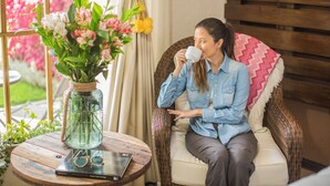 A person sitting on a wicker chair with a cushion, holding a cup, in a room with a wooden table, a vase with flowers, and a window with curtains. - Lamay Lodge by Mountain Lodges of Peru (Lamay)