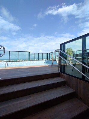 Zona de piscina moderna al aire libre con vista al mar, escalones de madera y barandillas de vidrio.
