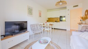 A modern living room with a flat-screen TV, a sofa, a dining area with a table and chairs, and a kitchen in the background.