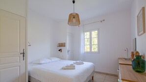 A bedroom with a bed, a window, a hanging light fixture, and a green vase on a wooden dresser.