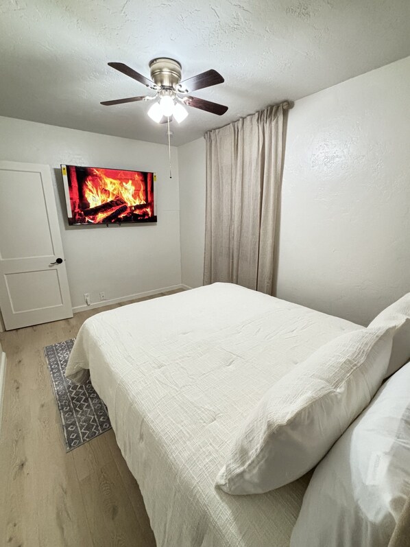Un dormitorio con cama, ventilador de techo, televisor mostrando una chimenea y una puerta.