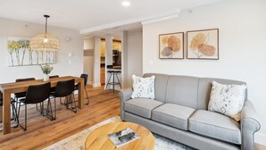 Una sala de estar moderna con un sofá gris, una mesa de comedor de madera con sillas, una lámpara colgante y dos cuadros enmarcados en la pared.