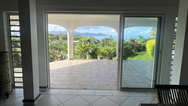 A spacious room with a large sliding glass door leading to an outdoor patio with a view of mountains and greenery. - Te Tai Marama (Raiatea)
