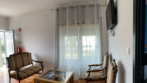 A living room with a wooden floor, two upholstered chairs, a glass coffee table, and a large window with sheer curtains. - Apartment 'Alojamento D Duarte T1' with Mountain View, Balcony and Wi-Fi (Gouveia)