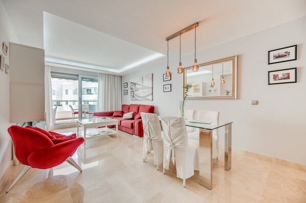 A modern living room with a red sofa, white chairs, a glass dining table, and a large mirror.