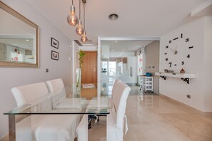 A modern living room with a glass dining table, white sofas, a large mirror, and a wall clock.