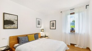 A bedroom with a wooden bed, a nightstand, a lamp, and a window with sheer curtains.