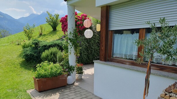 A house with a paved entrance, potted plants, a window with a curtain, and a view of a green landscape with mountains.