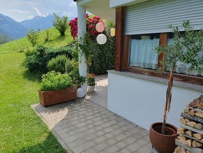 Una casa con entrada pavimentada, plantas en macetas, una ventana con cortina y vistas a un paisaje verde con montañas.