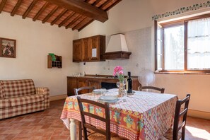 A dining area with a table set for two, wooden chairs, a checkered tablecloth, a fan, and a window with curtains.