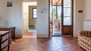Une porte ouverte donnant sur un balcon avec vue sur les arbres, un sol carrelé, un canapé à carreaux et un coin repas avec une table et des chaises.