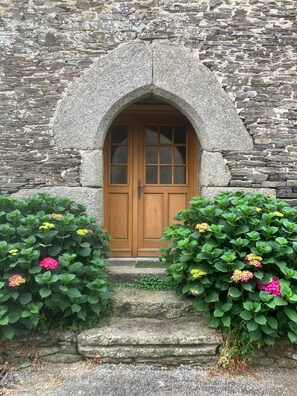 Un edificio de piedra con puerta de madera, un arco de piedra y plantas verdes con flores rosadas al frente.
