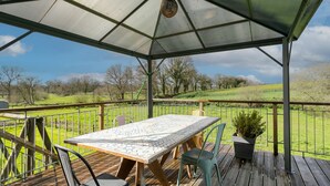 Eine überdachte Terrasse im Freien mit Tisch und Stühlen, einem Blumentopf und Blick auf ein grünes Feld.