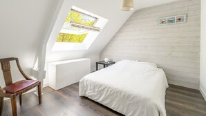 Quarto com teto inclinado, cama, poltrona, mesinha, abajur e janela com vista para árvores.
