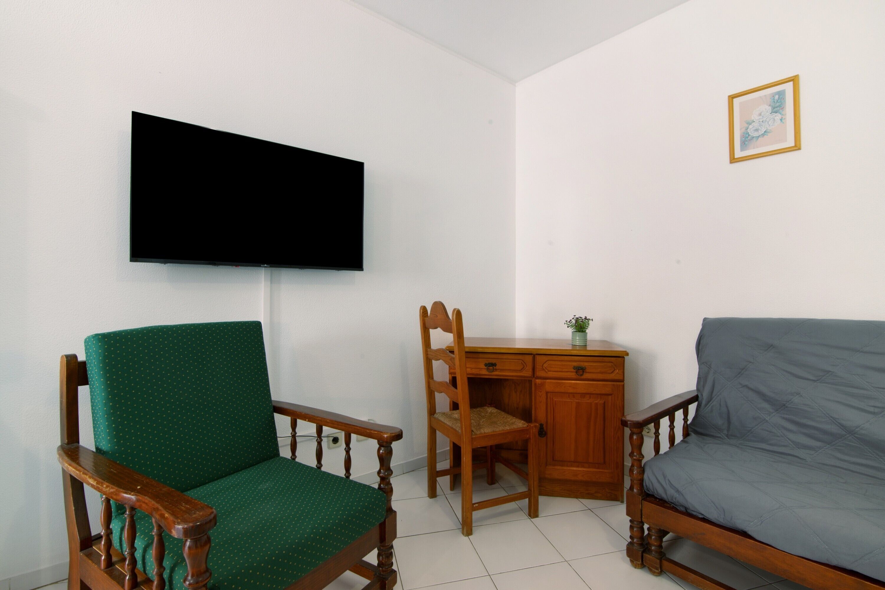 A room with a green upholstered chair, a wooden desk with a small plant, a grey sofa, and a flat-screen TV mounted on the wall.