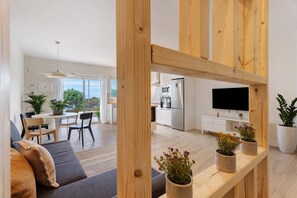 En moderne stue med en sofa, et træbord med plantepotte, et fjernsyn og et køkken i baggrunden.
