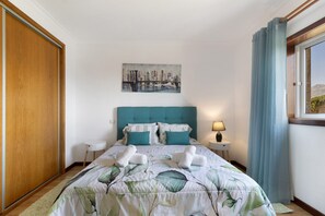Une chambre d’hôtel avec un lit, un tableau représentant un paysage urbain, une fenêtre avec des rideaux et une table de chevet avec une lampe.