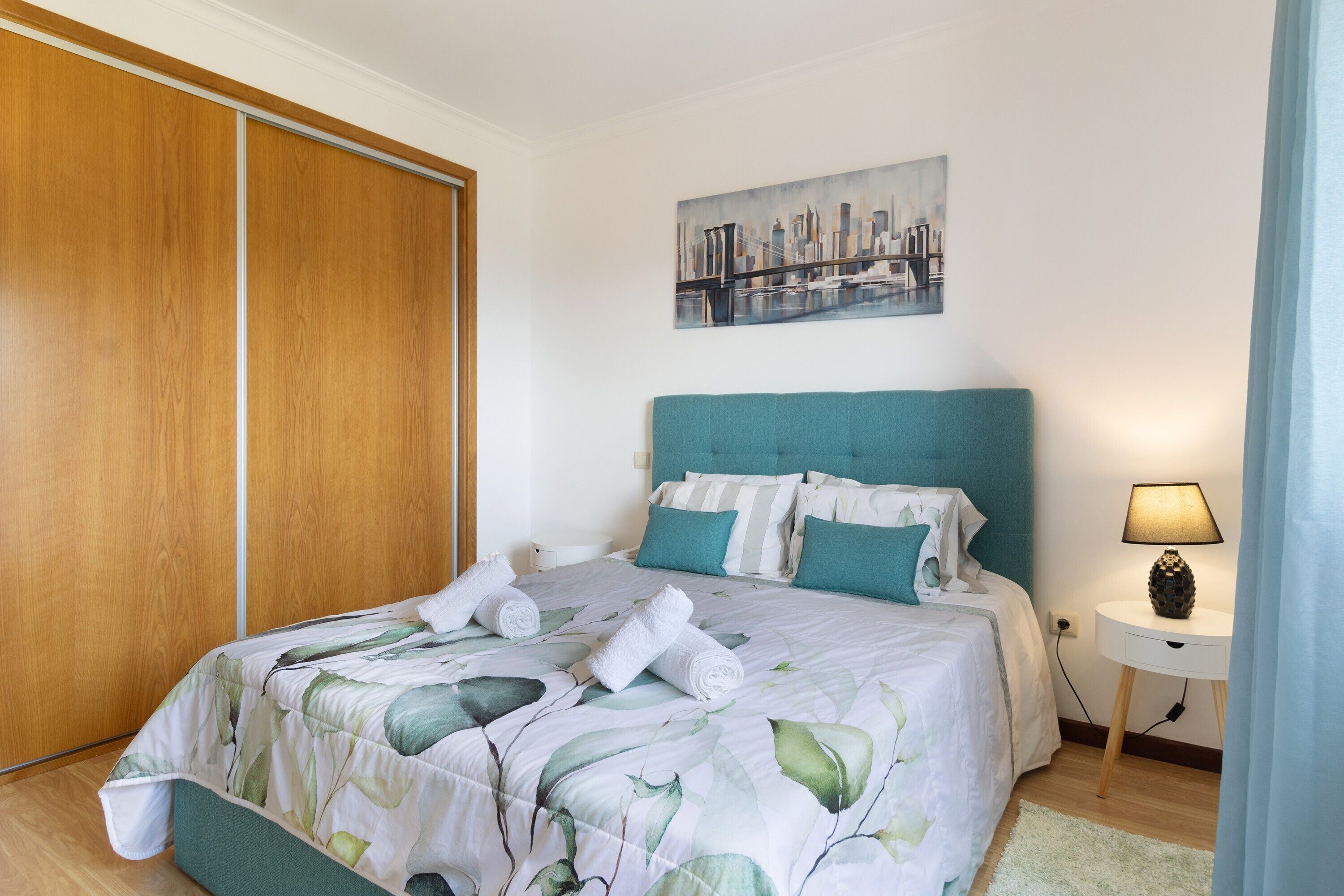Ein ordentlich bezogenes Bett mit Blumenmuster, ein Stadtbild über dem Bett, ein Holzschrank und eine Nachttischlampe.