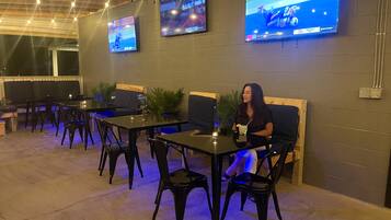 Un espace extérieur aménagé avec des tables et des chaises, un téléviseur fixé au mur et des guirlandes lumineuses suspendues au plafond.