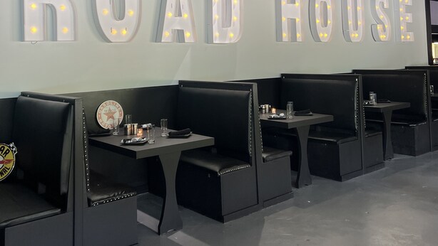 L’intérieur d’un restaurant, avec une enseigne lumineuse « ROAD HOUSE », des banquettes noires et des tables avec des verres et des condiments.