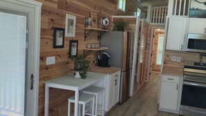A compact kitchen with wooden walls, white cabinetry, a refrigerator, a microwave, an oven, and a small dining area with a table and benches. - Rustic Tiny Home Escape - Pets Stay Free! (Georgetown)