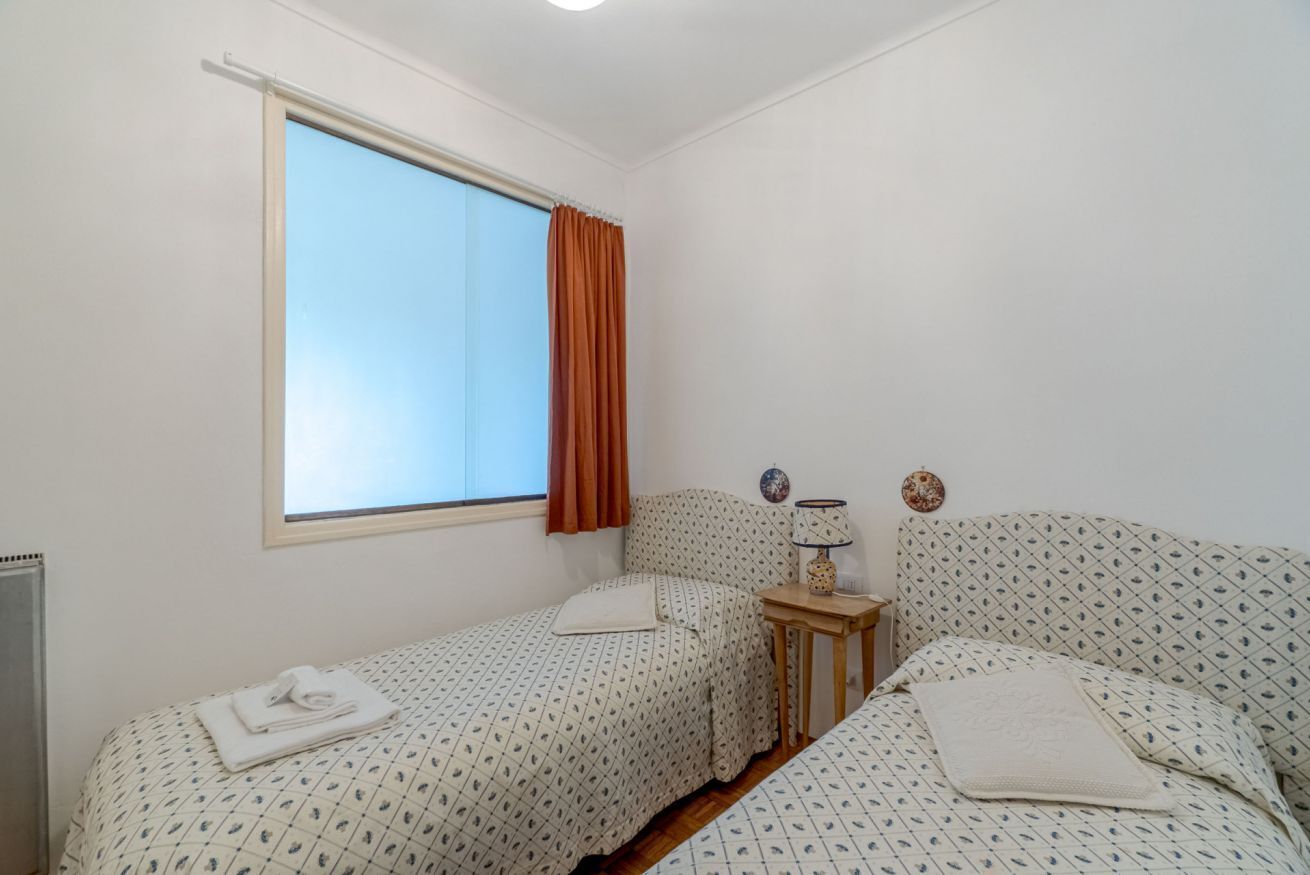 Una habitación pequeña y limpia con dos camas, una ventana con cortinas, una mesita de noche con lámpara y un radiador.