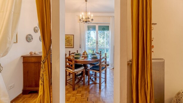 Un comedor con mesa y sillas de madera, una araña y vistas a través de un ventanal.