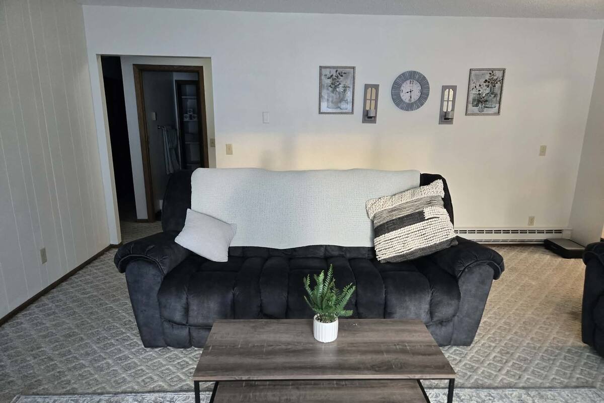 Un salon avec un canapé noir, une table basse en bois et une plante en pot. On y trouve des horloges murales et des tableaux encadrés accrochés au mur.