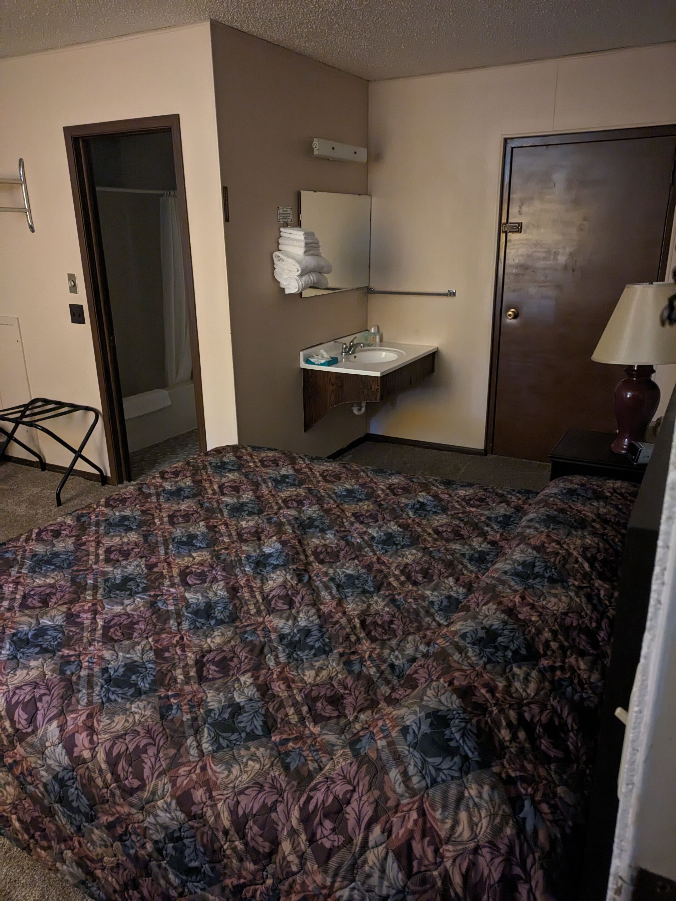 Une chambre d’hôtel avec une couvre-lit à motifs floraux, une salle de bain avec un lavabo et un miroir, et une porte sur le côté droit.