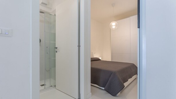A modern bedroom with a glass shower, a bed with a grey blanket, and a white wall with a light fixture. - GuestHost - Academy of Fine Arts -Modern Apt. with balcony! (Bologna)