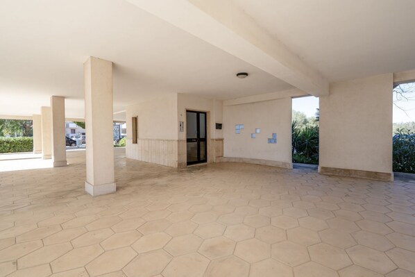 Un espacio amplio y cubierto con suelo de baldosas hexagonales, columnas y una puerta que da a un espacio interior.