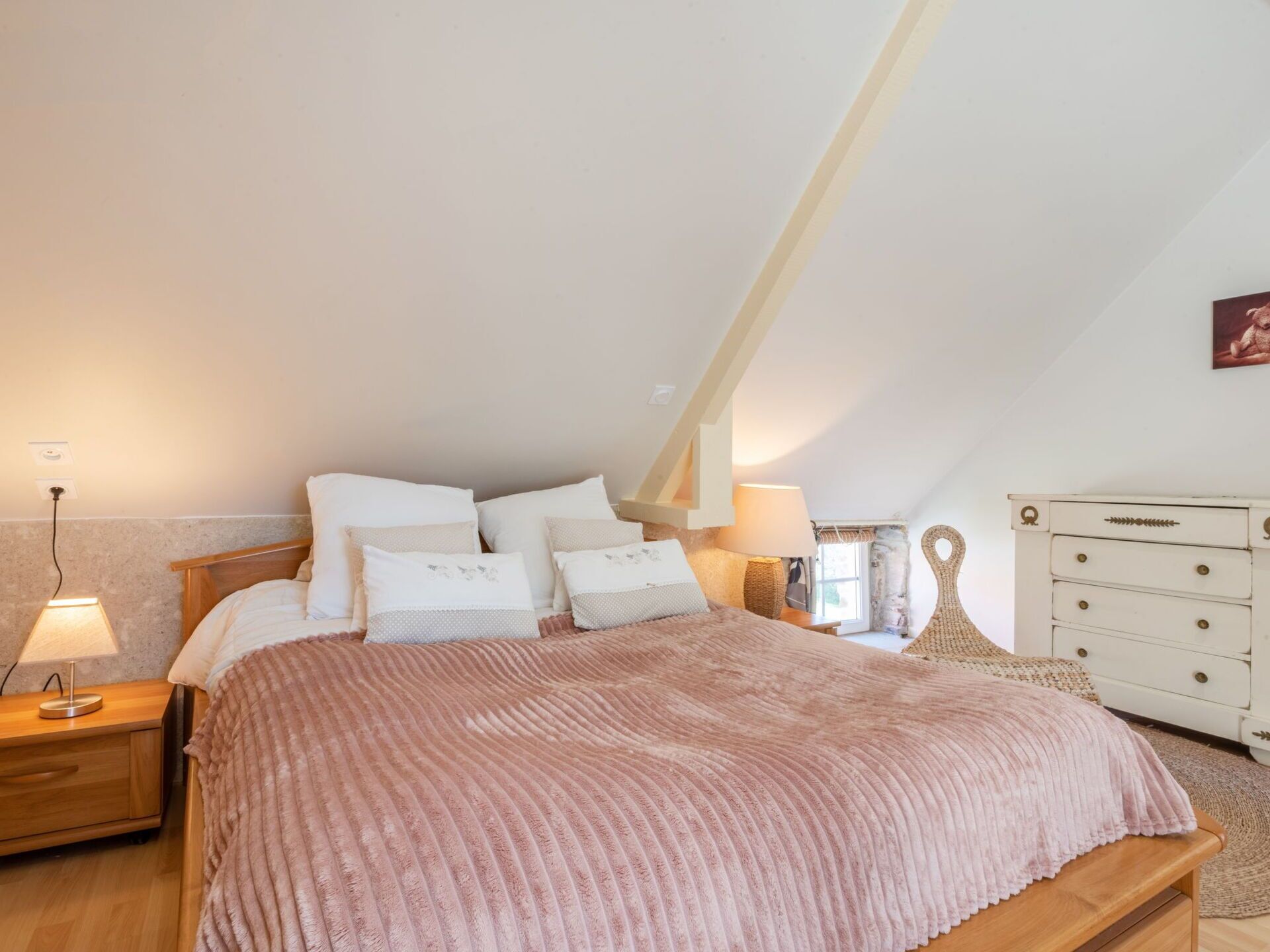 Ein Schlafzimmer mit geneigter Decke, einem hölzernen Bett mit rosa Decke, weißen Kissen, einem Nachttisch mit Lampe und einer weißen Kommode mit Schubladen.